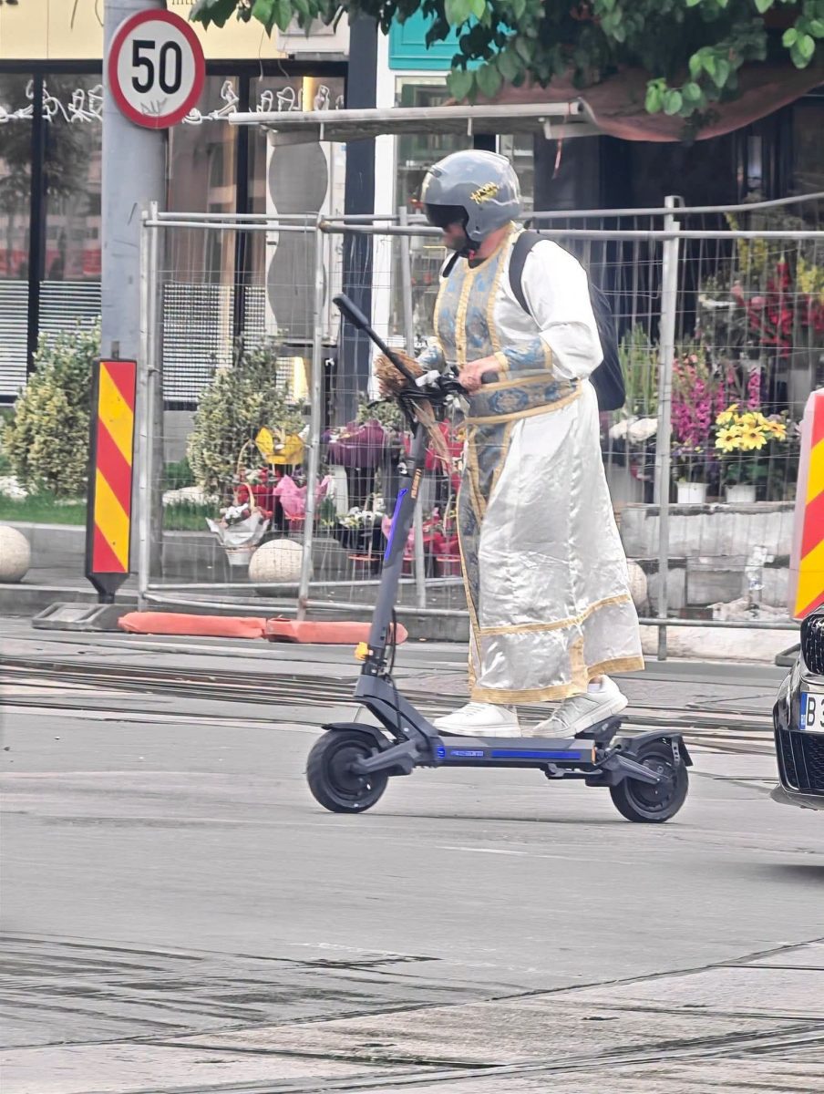 Osoba v oblečení jazdí na elektrickej kolobežke ulicou, obklopená kvetmi a stavebným zátiším.