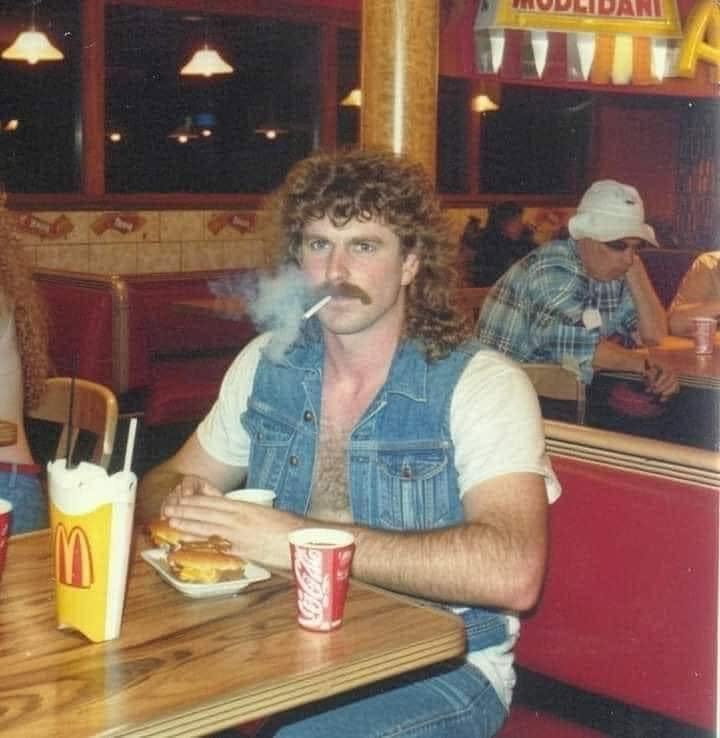 Muž s kučeravými vlasmi a cigaretou v reštaurácii, sedí pri stole s jedlom a nápojom. Retro atmosféra 80. rokov.