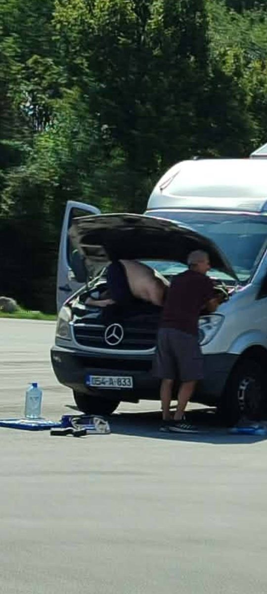 Muž opravuje motor dodávky na parkovisku, pričom jeden z nich je hlavou dolu v motorovom priestore.