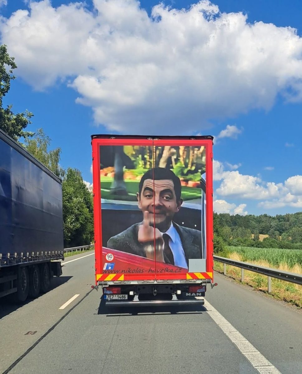 Nákladné auto s obrázkom známeho komika na zadných dverách jazdí po vidieckej ceste za slnečného dňa.