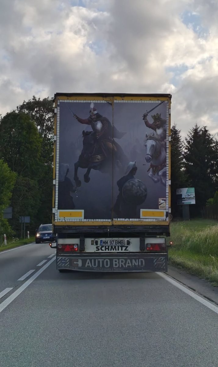 Kamión s historickou maľbou rytierov na zadnej strane, jazdiaci po vidieckej ceste pod zamračenou oblohou.