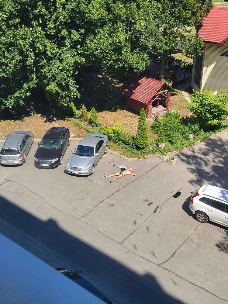 Osoba opaľujúca sa na parkovisku v blízkosti áut a zelene počas slnečného dňa.