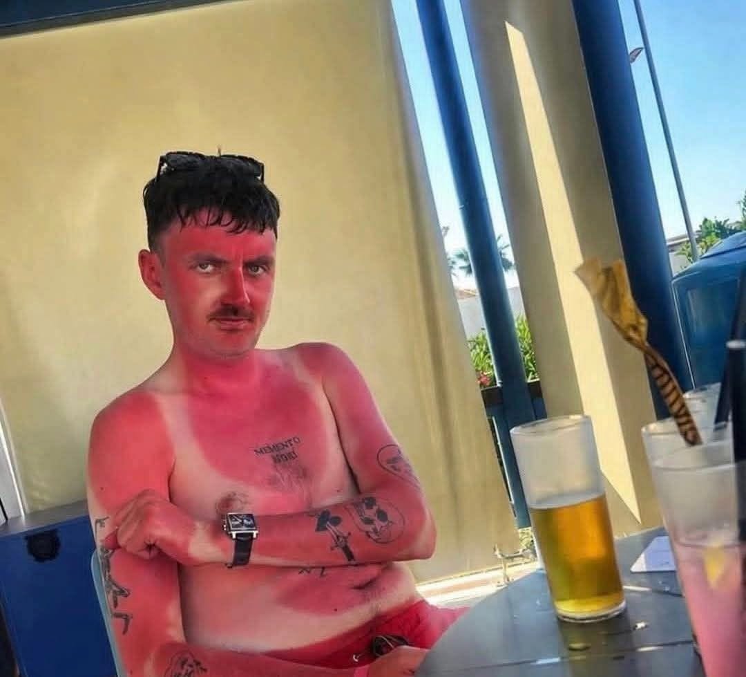 Muž so spálenou pokožkou sedí pri stole na terase, vedľa nápojov a s tetovaniami na rukách.