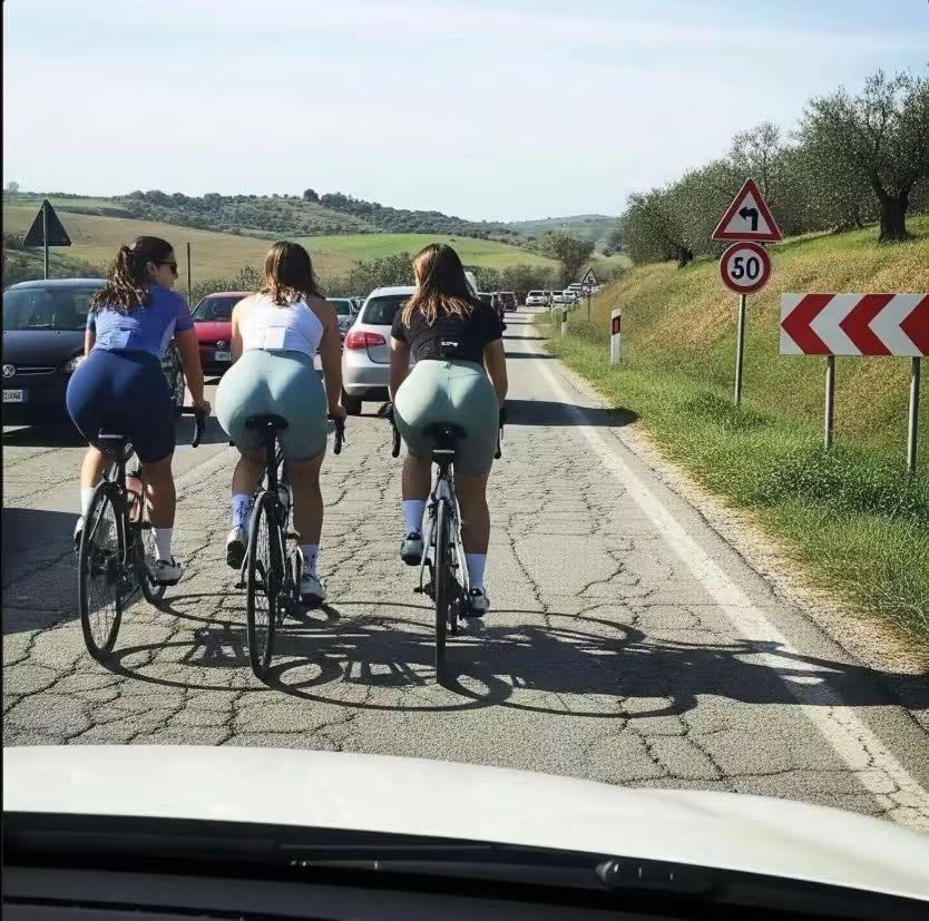 Tri cyklistky na vidieckej ceste, jazda vedľa seba, obklopené autami a krajinkou s dopravnými značkami.