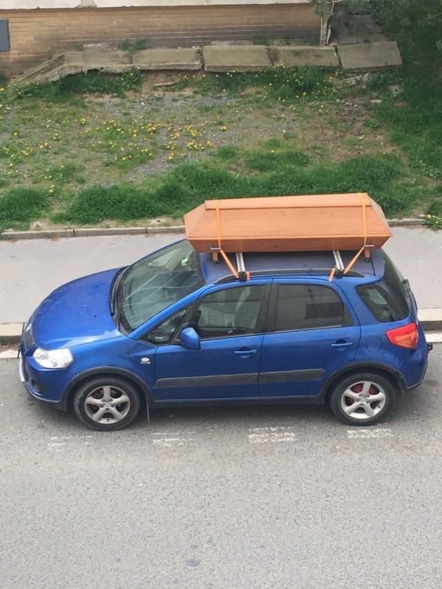 Modré auto s drevenou rakvou upevnenou na streche, zaparkované na ulici.