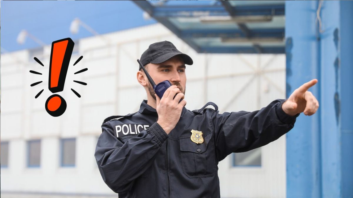 Policajt komunikuje cez vysielačku a ukazuje smer vonku, v pozadí moderná budova.