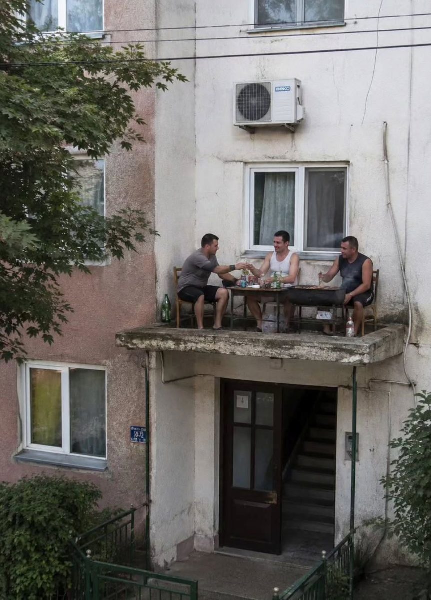 Traja muži si užívajú posedenie na balkóne nad vchodom budovy, obklopení prírodnými prvkami a mestskou architektúrou.