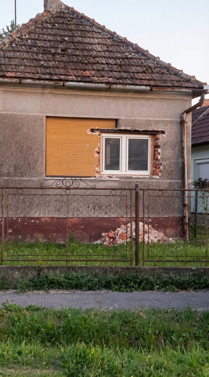 Starý dom s opravovaným oknom a nahromadenými tehlami, zelená tráva a staré kovové ploty.