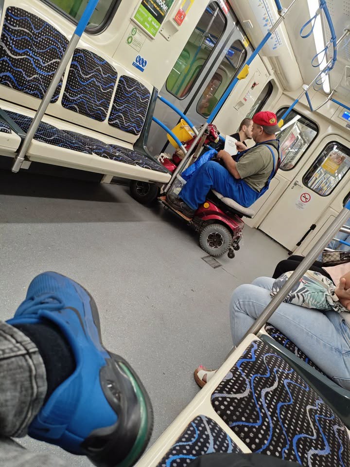 Muž na invalidnom vozíku v metre, čítajúci list, obklopený prázdnymi sedadlami.