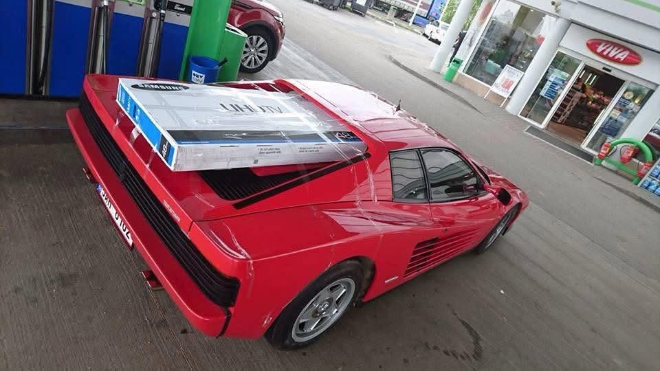 Červené športové auto s televízorom na kapote na čerpacej stanici.