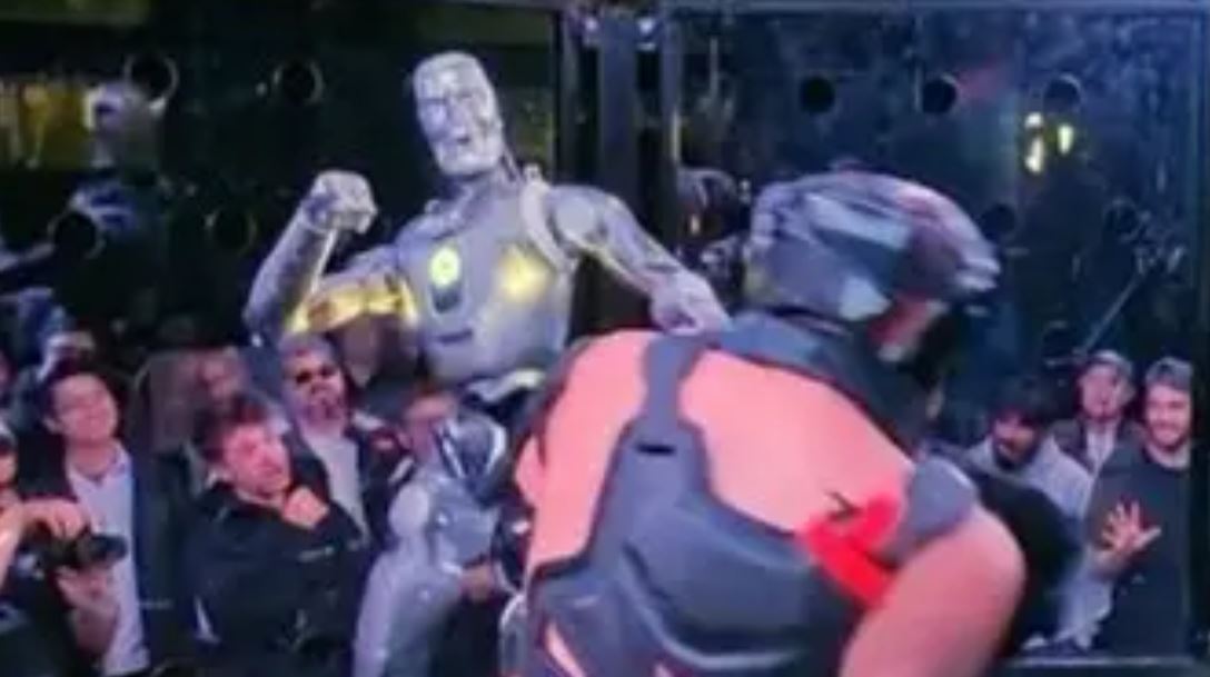 Robot a človek v ringu bojujú pred divákmi. Zápas technológie proti sile.
