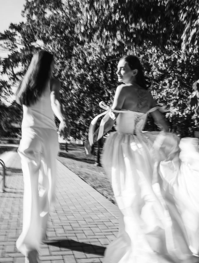 Dve ženy kráčajú v parku, jedna v svadobných šatách, čiernobiela fotografia, letné prostredie.