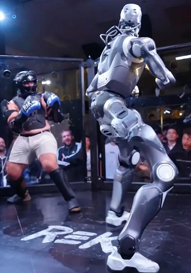 Robot bojuje v ringu proti človeku v ochrannom oblečení počas zápasu v aréne.