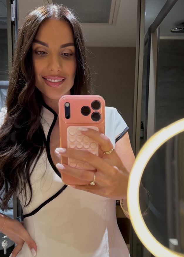 Osoba v zrkadle si robí selfie; má dlhé hnedé vlasy a biely outfit, s mobilom v ružovom kryte.