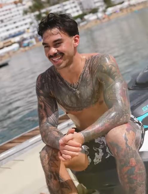 Muž s tetovaním na pláži sedí na vodnom skútri, usmieva sa a relaxuje počas letného dňa.