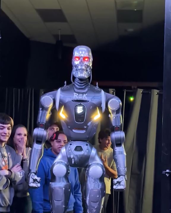 Robot s červenými očami obklopený skupinou ľudí v tmavej miestnosti.