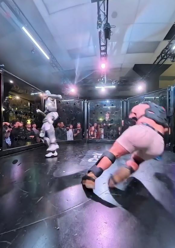 Robotický zápas na pódiu, kde dvaja bojovníci zápasia pred publikom obklopení osvetlením a kamerami.