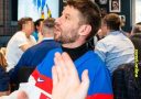 Muž v slovenskom drese s prekvapeným výrazom na skupinovom stretnutí ľudí v interiéri.