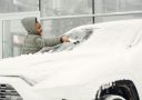 A woman in a gray coat clears snow from a car windshield on a cold winter day.