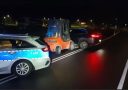 Policajné auto a vysokozdvižný vozík blokujú čierne auto na nočnej ceste.