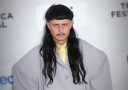 Osoba s dlhými tmavými vlasmi v šedom saku na červenom koberci festivalu Tribeca.