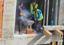 Robotníci na stavbe pripravujú obed na provizórnom grile z fúrika, obklopení stavebným materiálom.