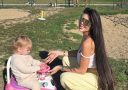 Žena v parku so slnečnými okuliarmi, kľačiaca vedľa dieťaťa na ružovom odrážadle.
